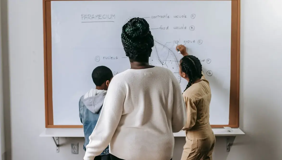 Professora escrevendo na lousa enquanto alunos acompanham a explicação